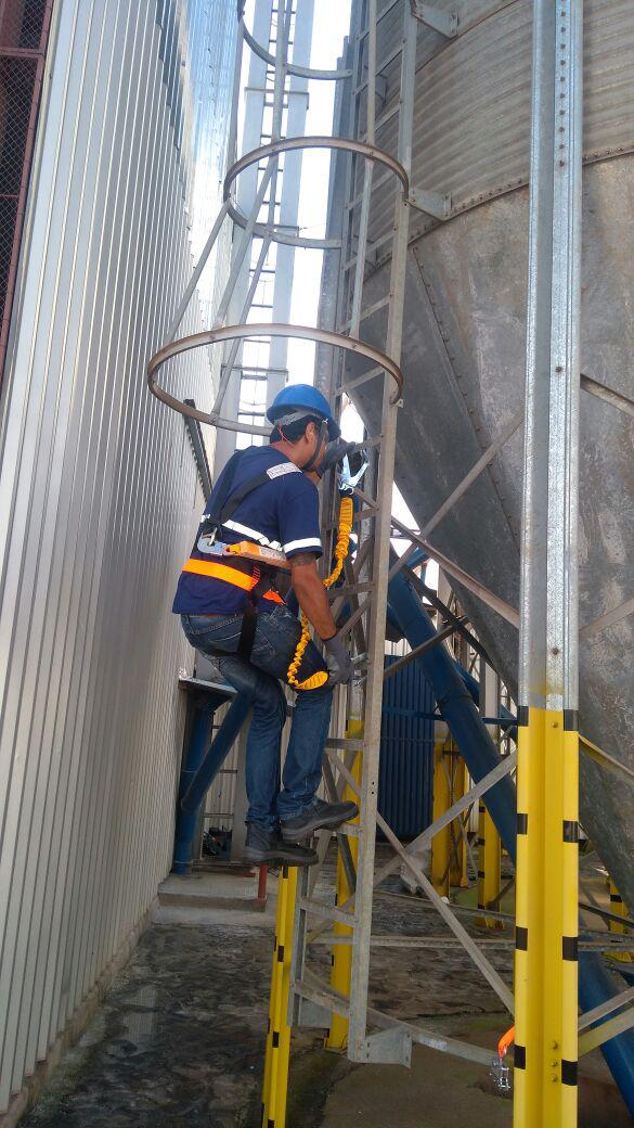 Operador com cinto de segurança e capacete, subindo escada tipo gaiola em estrutura industrial alta.